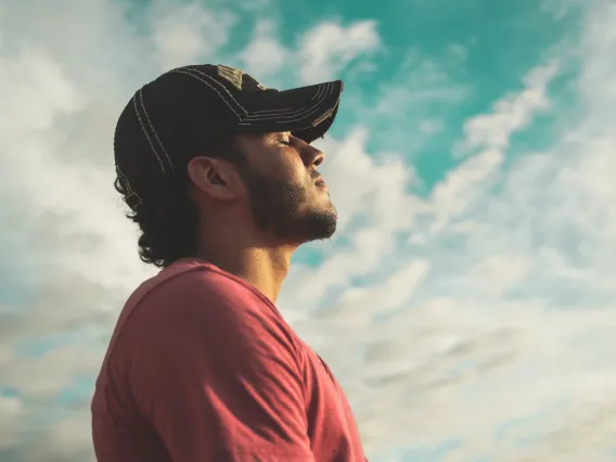 man wearing black cap with eyes closed under cloudy sky