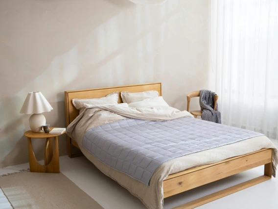 Tan wood bed with light blue sheets and off white pillow and bed stand with lamp.