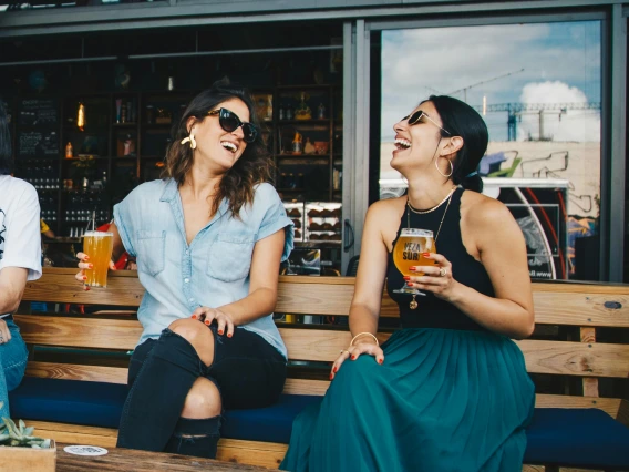 two women sitting and talking on a patio restaurant bench holding a beer