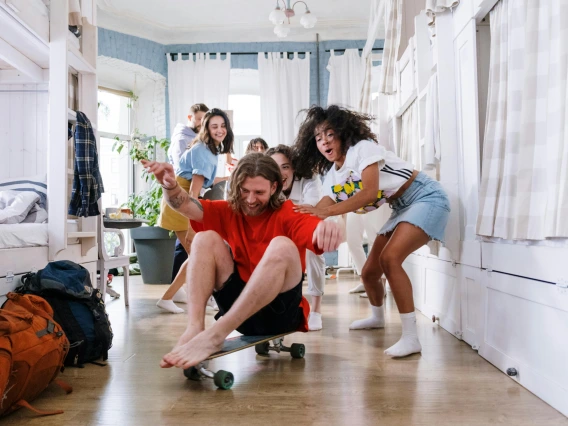 group of people laughing playing with a skateboard in a dorm room