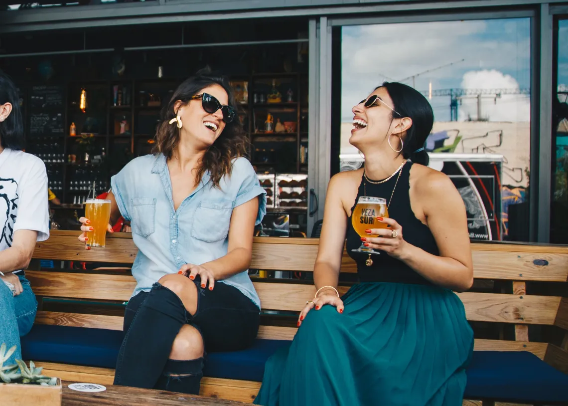 Two people sitting on a bench laughing and holding a beer