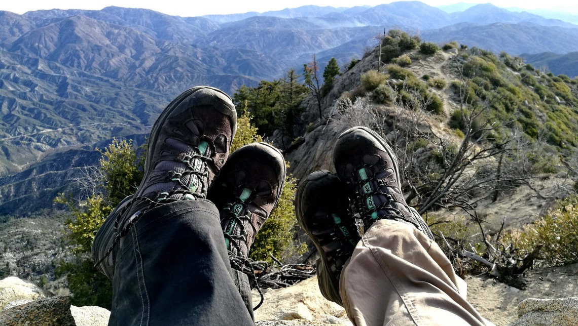Two people's feet with hiking boots on and mountains in the background