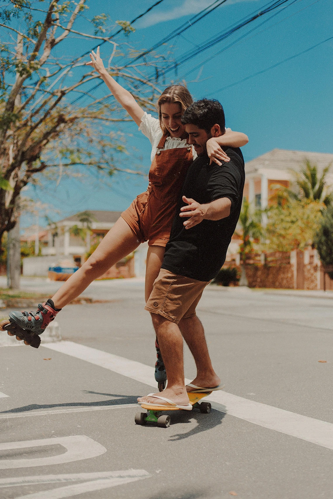 Person with black shirt and khaki shorts on skateboard holding other person on roller skates in overalls.